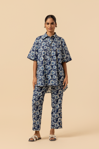 A model in a blue floral-patterned outfit stands against a soft beige background, showcasing a relaxed style.