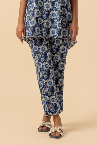 A person stands wearing  in a blue floral-patterned outfit, set against a soft beige background against a neutral backdrop.