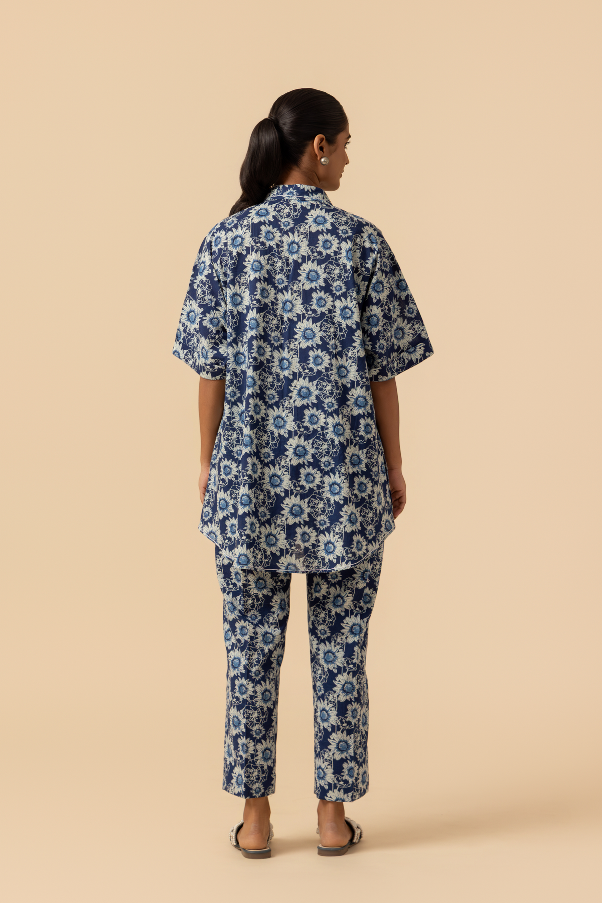 Back view of a model in a blue floral-patterned outfit stands against a soft beige background, showcasing a relaxed style.