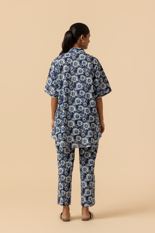 Back view of a model in a blue floral-patterned outfit stands against a soft beige background, showcasing a relaxed style.