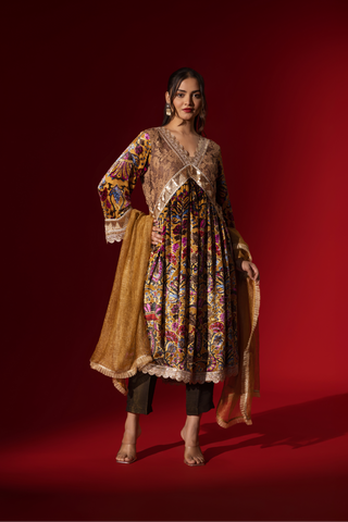 A woman stands confidently in a colorful, embellished regal printed velvet kurta set with a flowing golden dupatta against a vibrant red backdrop.