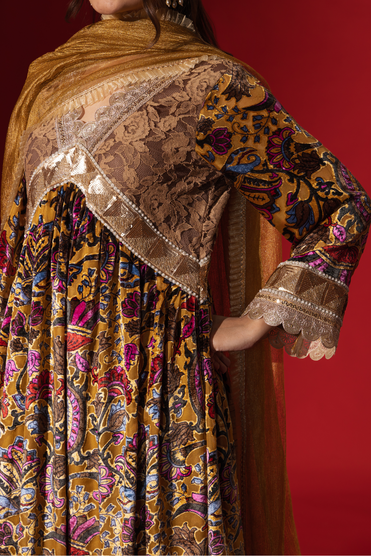 Closeup view of a woman's hand in a colorful, embellished regal printed velvet kurta set with a flowing golden dupatta against a vibrant red backdrop.