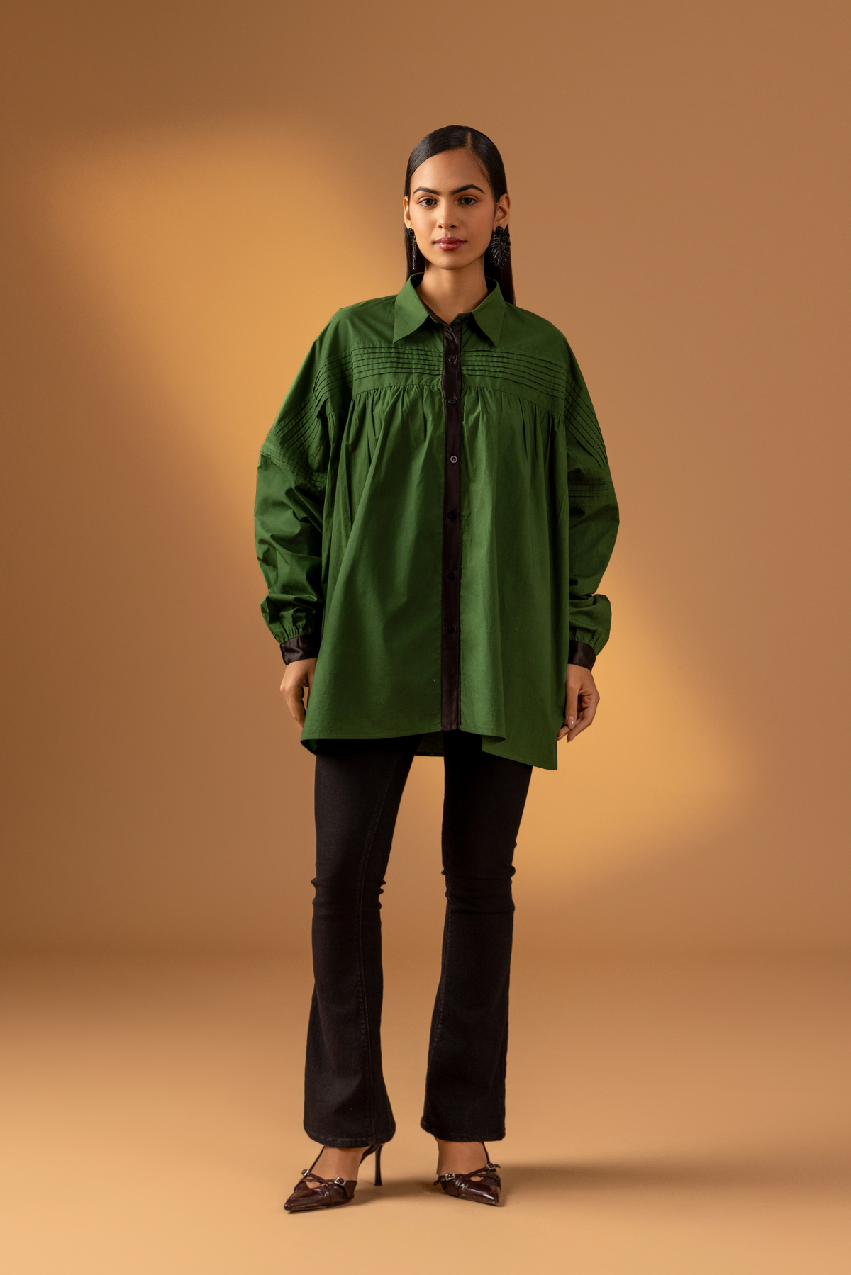 A model stands against a warm beige background, wearing a loose green shirt with a black button front and black flared pants .