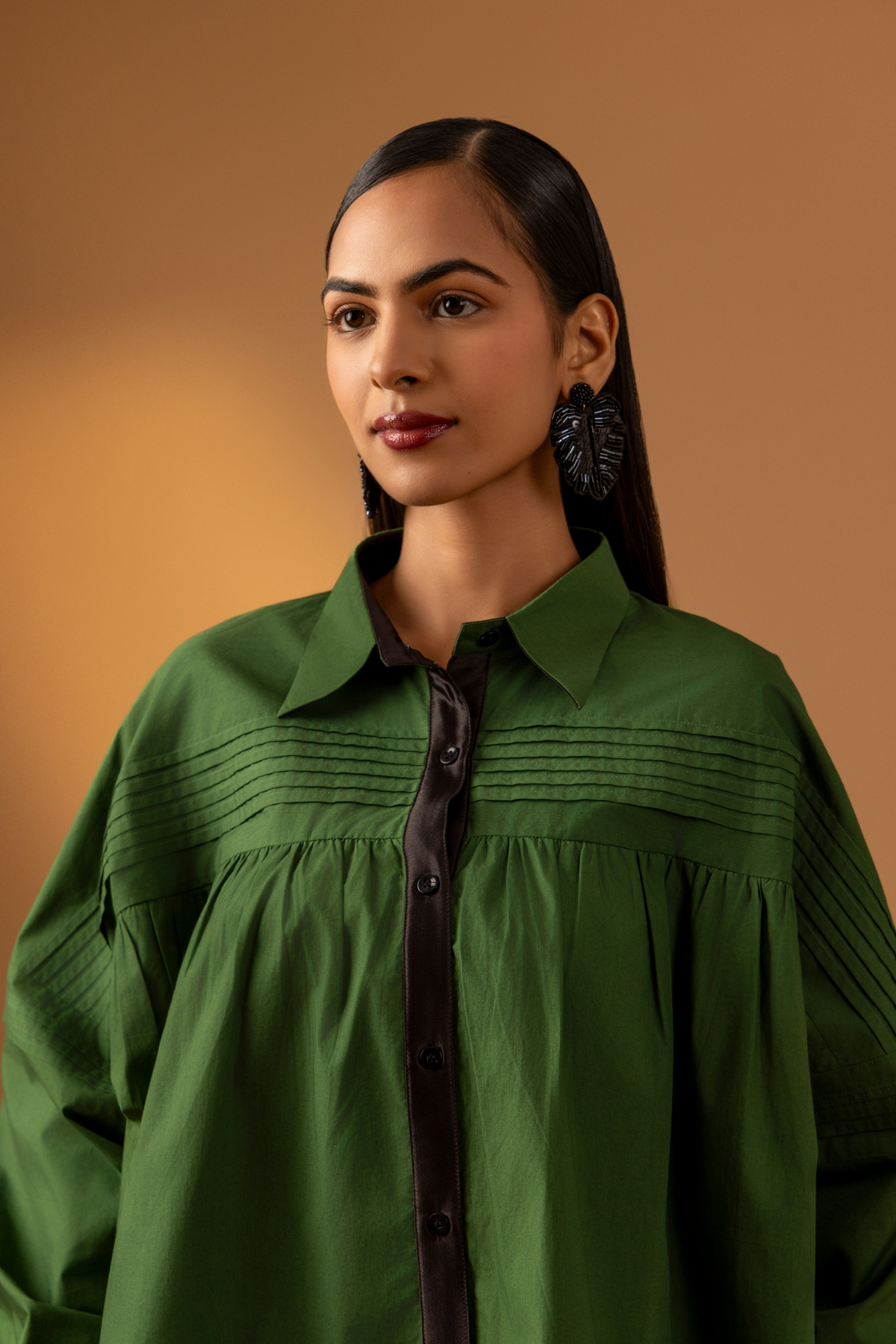 Closeup view of a woman stands against a warm beige background, wearing a loose green shirt with a black button front and black flared pants .