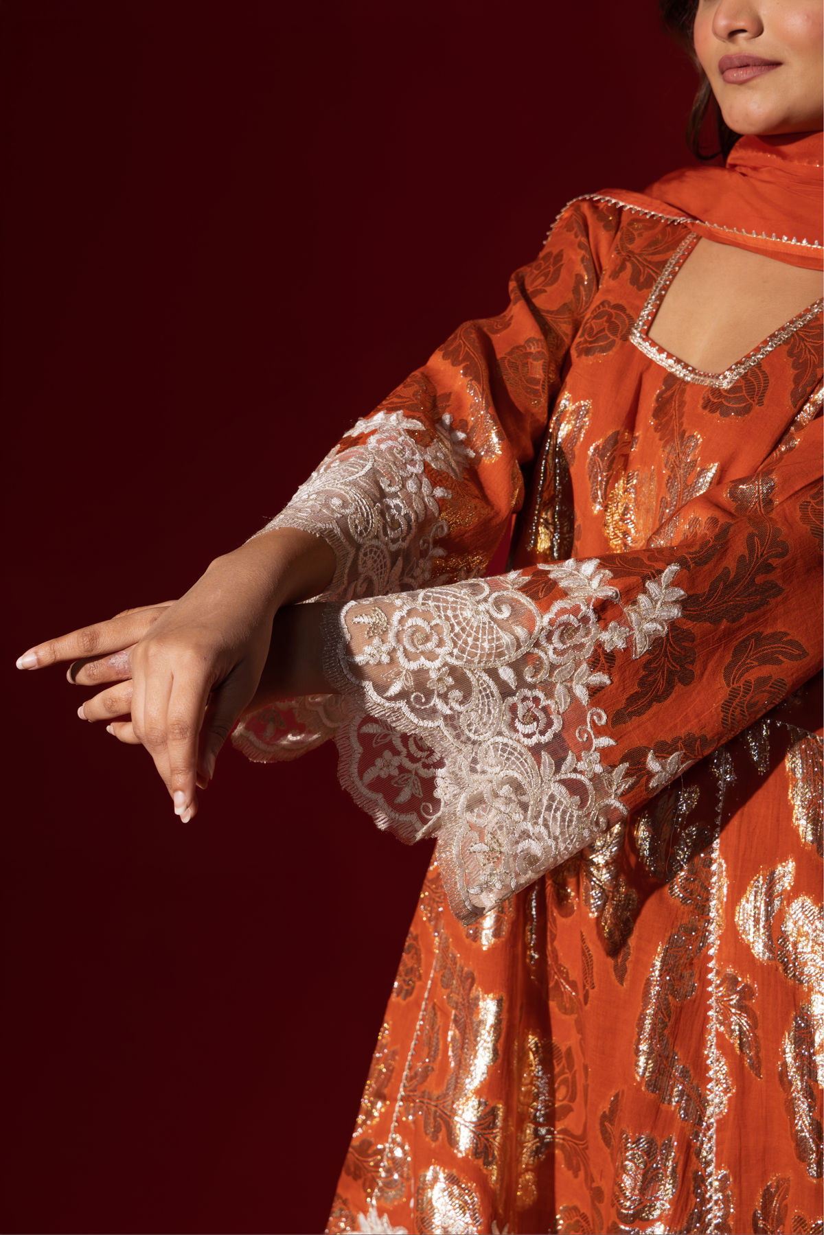 Closeup view of a woman 's hand in a orange ethnic  shimmer kurta set outfit with intricate white embroidery against a vibrant red backdrop.