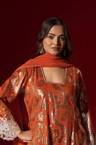 Closeup view of a woman poses in a orange ethnic  shimmer kurta set outfit with intricate white embroidery against a vibrant red backdrop.