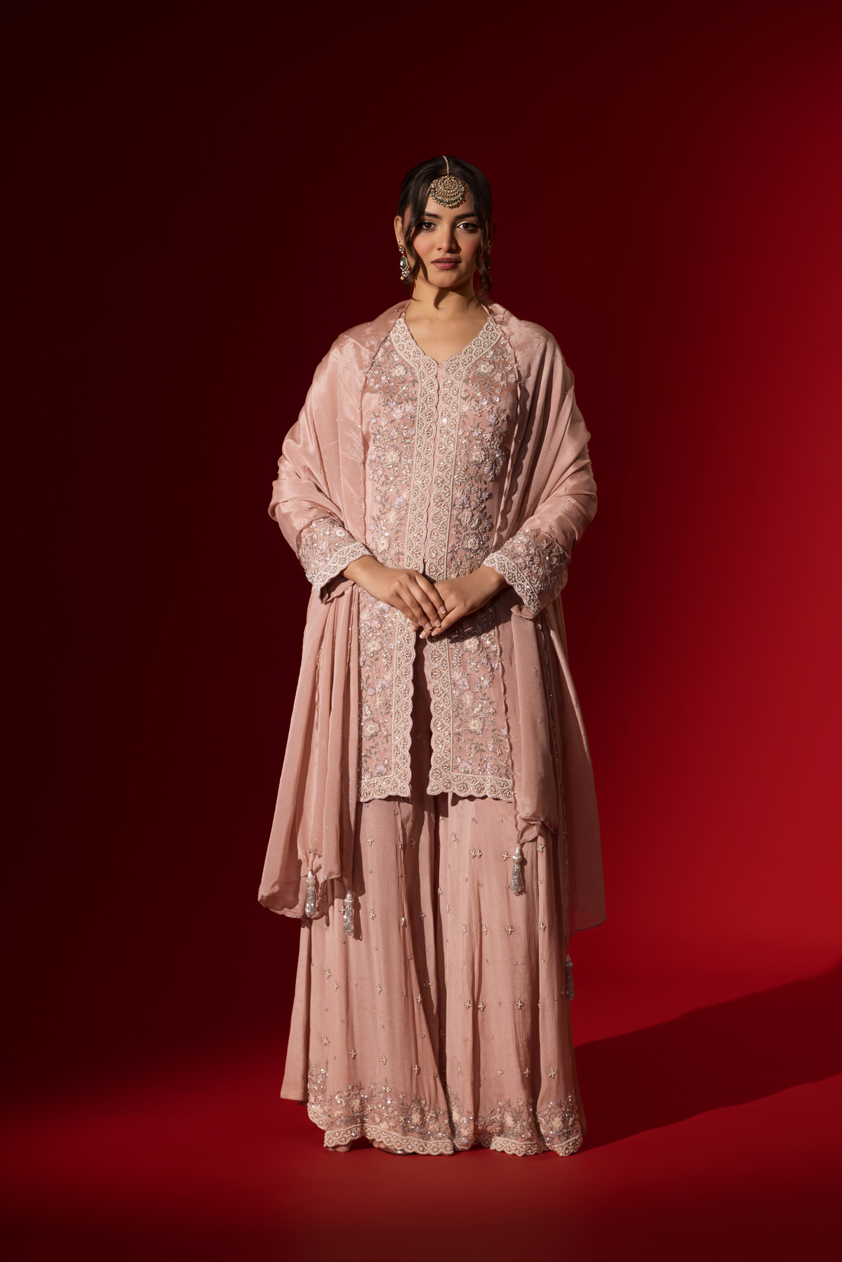 A woman wears an elegant pastel pink embroidered kurta set with intricate beadwork, standing against a vibrant red background.