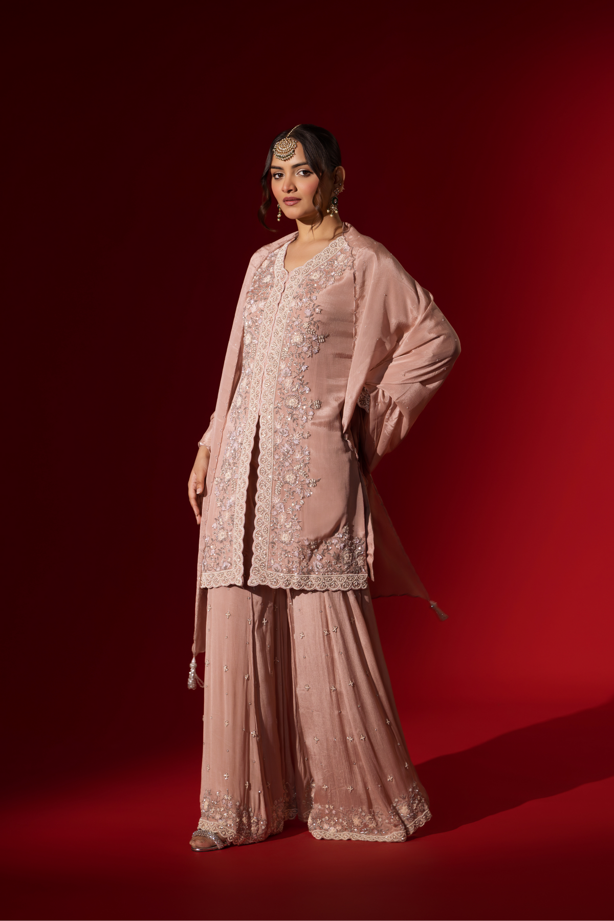 Side view of a woman wears an elegant pastel pink embroidered kurta set with intricate beadwork, standing against a vibrant red background.