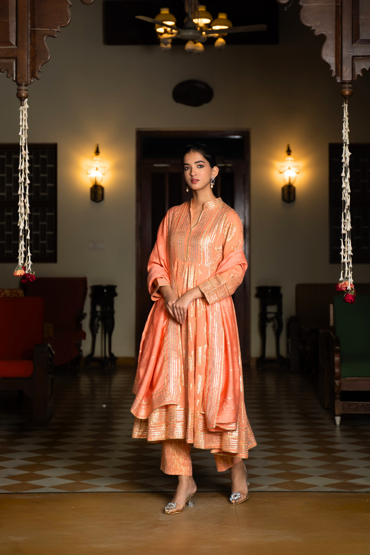 Side view of a  woman stands elegantly in an orange, traditional anarkali kurta set showcasing intricate embroidery against a beautifully decorated interior.