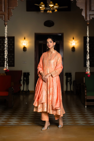 Side view of a  woman stands elegantly in an orange, traditional anarkali kurta set showcasing intricate embroidery against a beautifully decorated interior.