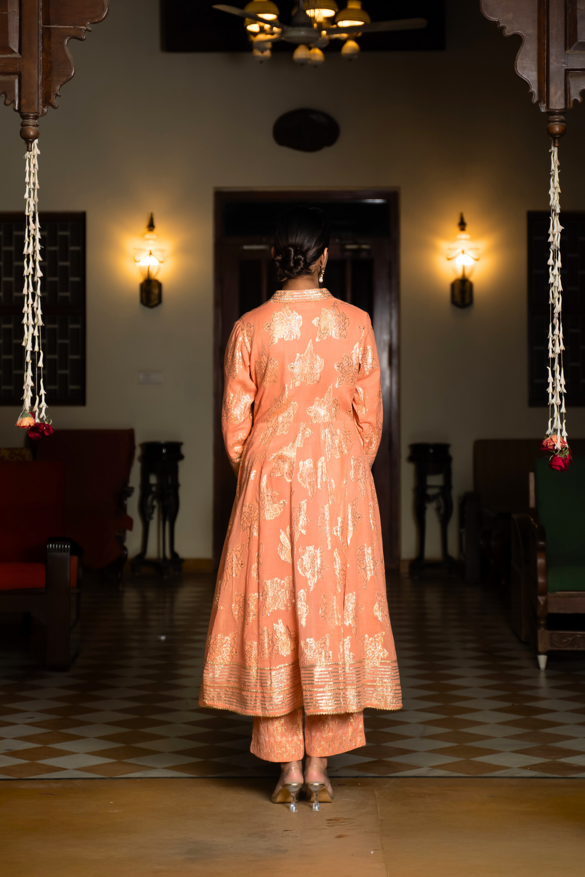 Back view of a  woman stands elegantly in an orange, traditional anarkali kurta set showcasing intricate embroidery against a beautifully decorated interior.