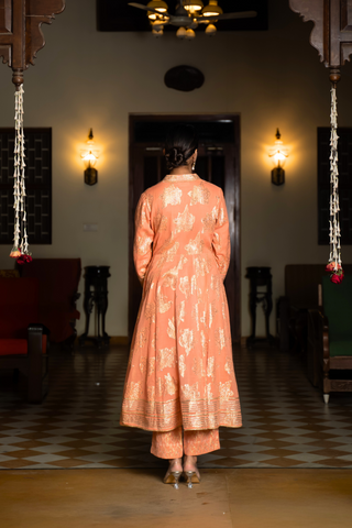 Back view of a  woman stands elegantly in an orange, traditional anarkali kurta set showcasing intricate embroidery against a beautifully decorated interior.