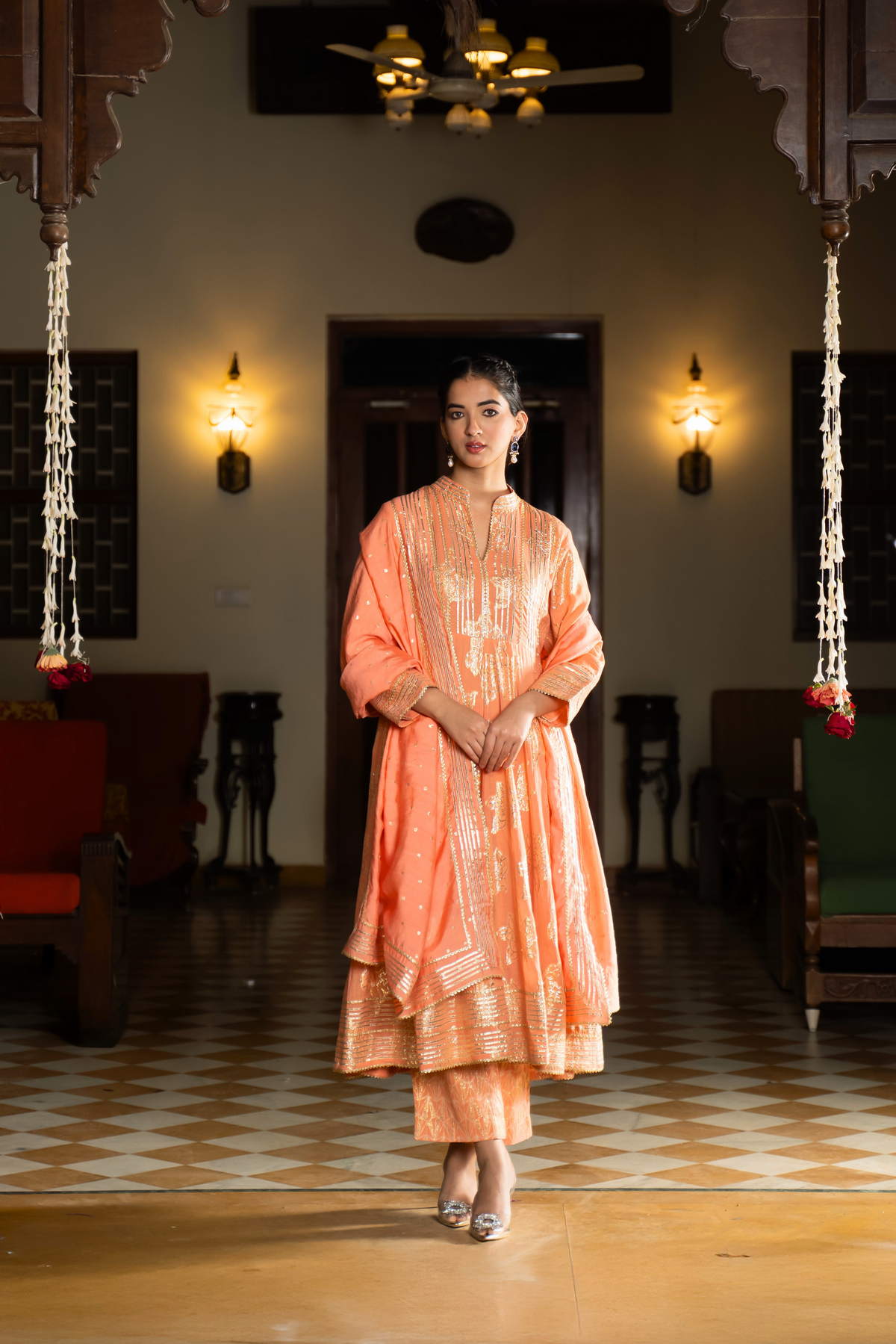 A person stands elegantly in an orange, traditional anarkali kurta set showcasing intricate embroidery against a beautifully decorated interior.