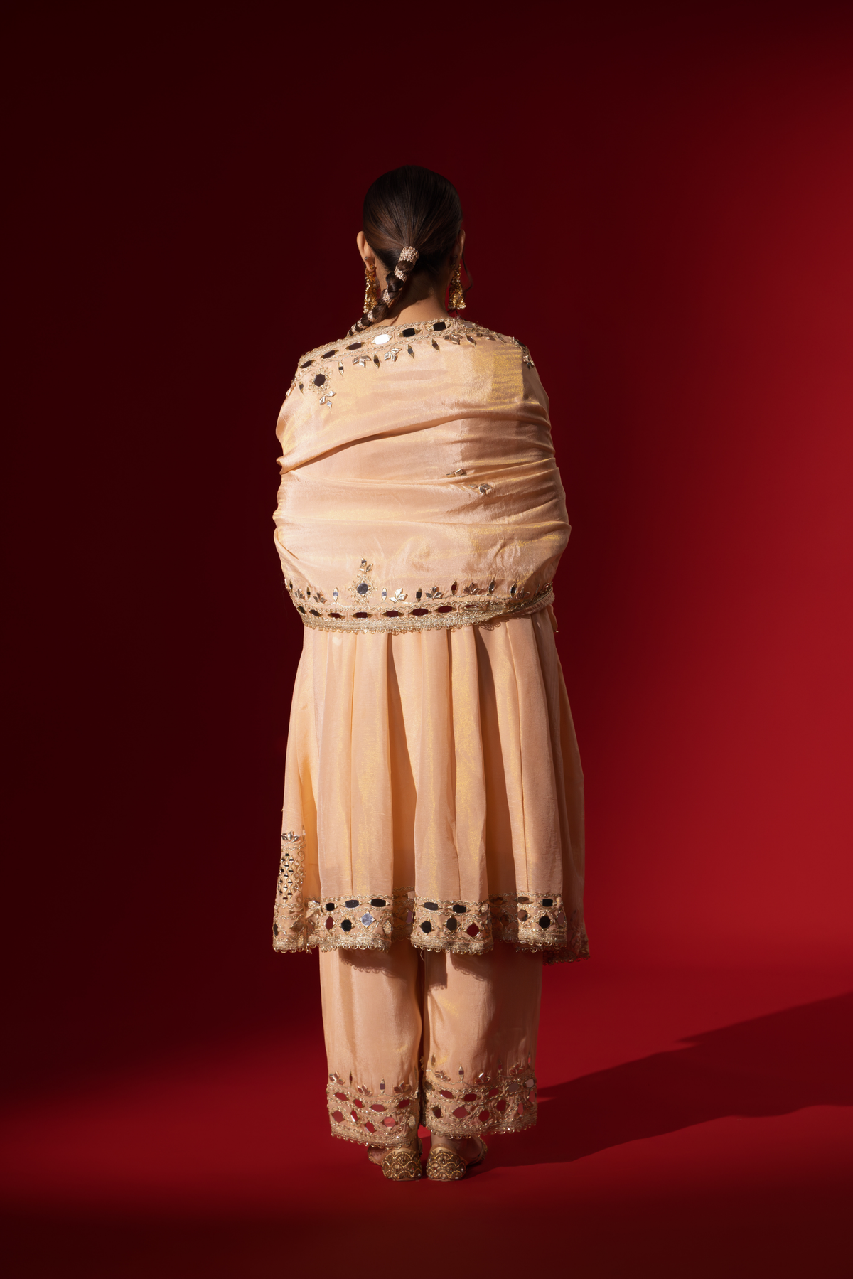 Back view of a woman stands elegantly in a beige embroidered kurta set with intricate patterns, complemented by a matching dupatta against a bold red background.
