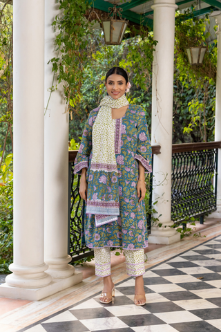 A woman wears a colorful, peacock-printed cotton kurta set with a matching scarf, standing on a checkered floor surrounded by greenery.