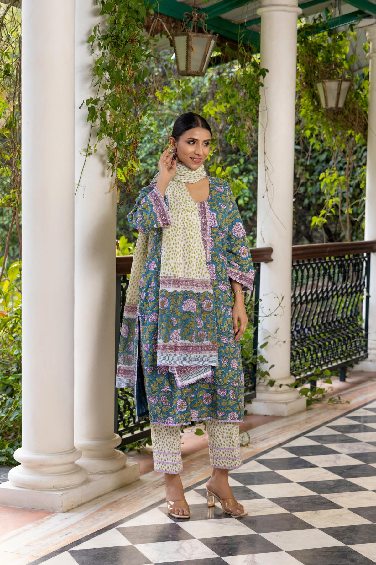 Right side view of  a woman wears a colorful, peacock-printed cotton kurta set with a matching scarf, standing on a checkered floor surrounded by greenery.