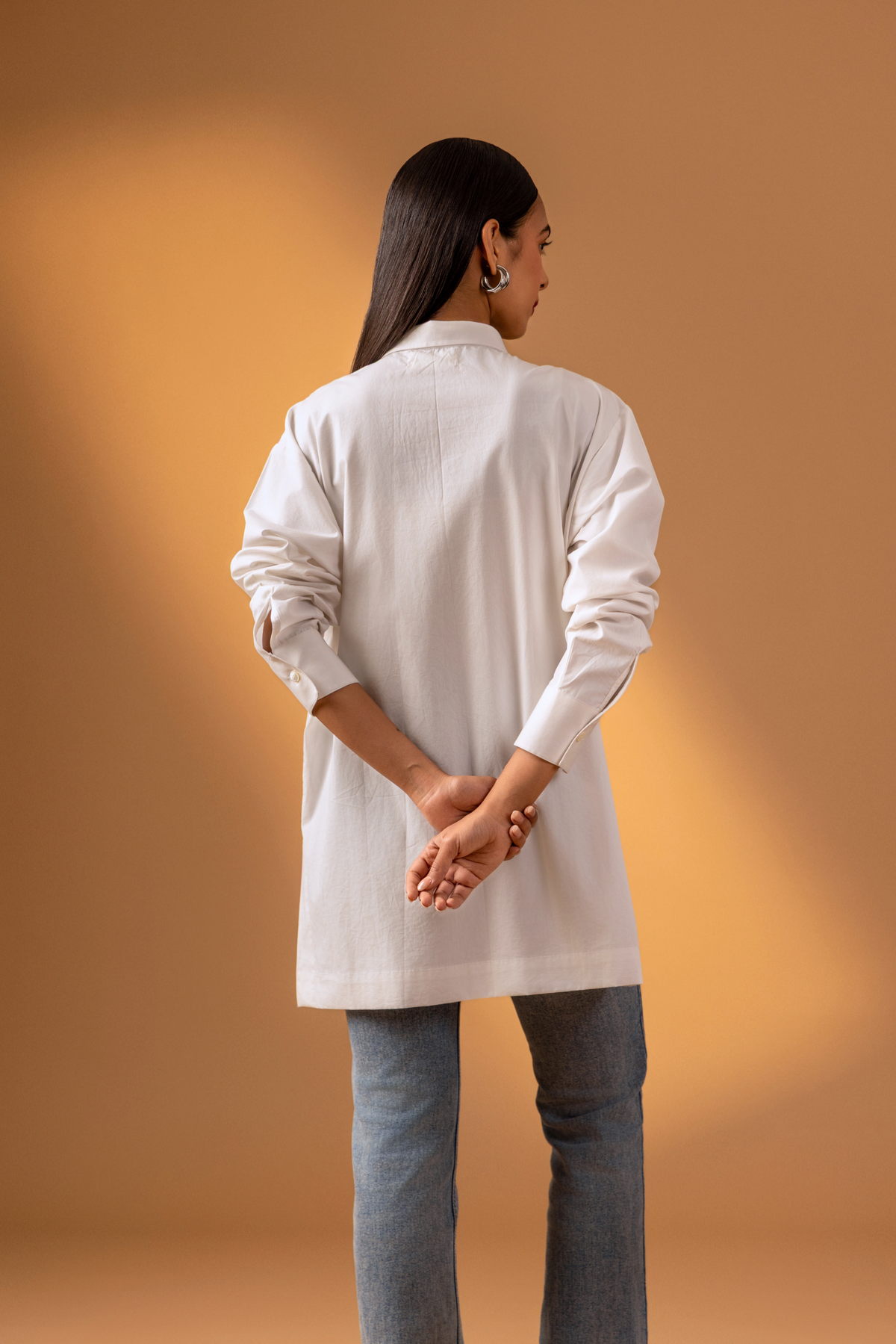Back view of a woman is dressed in a stylish white button-up shirt with long sleeves, paired with denim jeans, set against a warm beige background.