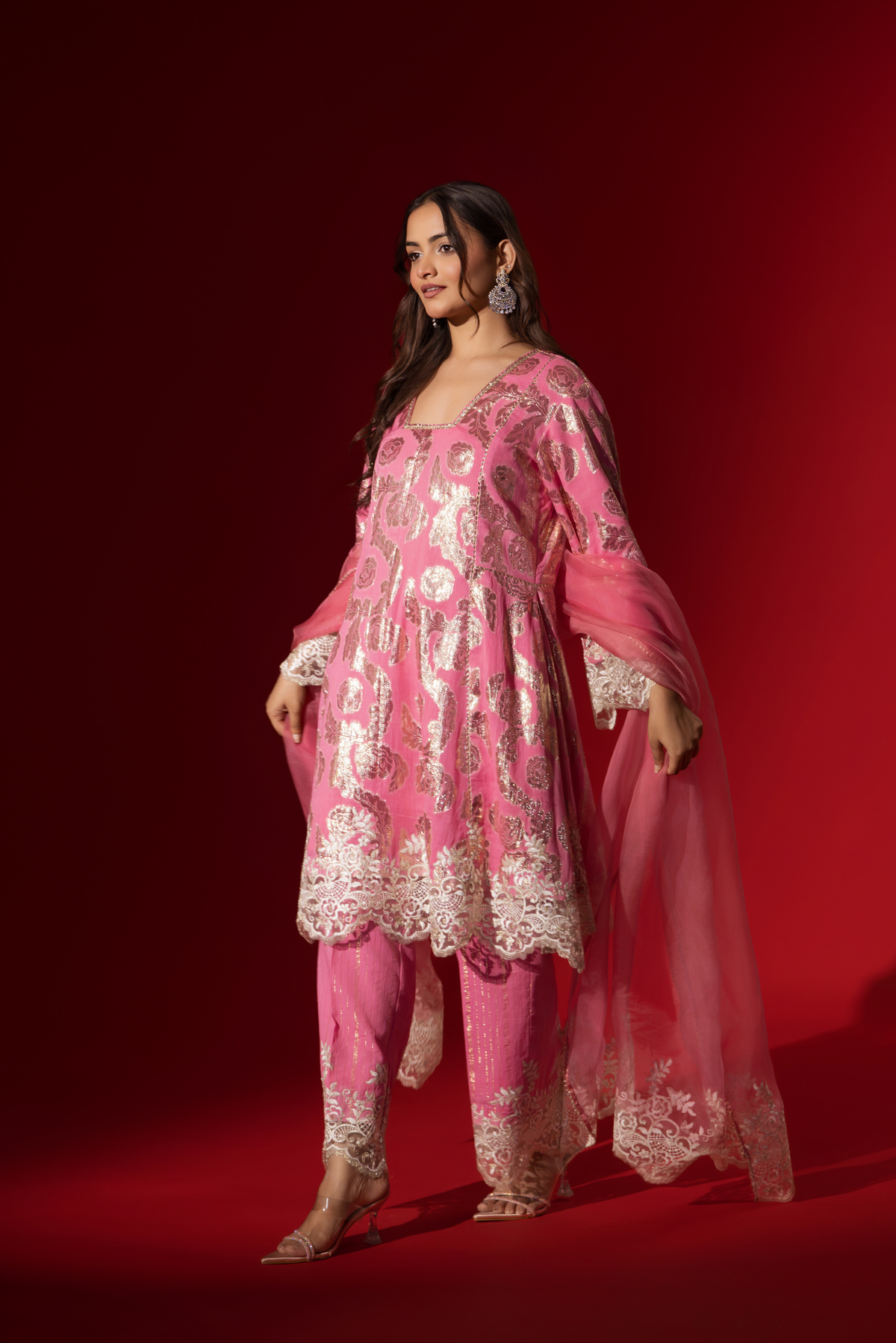 Side view of a woman poses in a pink ethnic shimmer kurta set outfit with intricate white embroidery against a vibrant red backdrop.