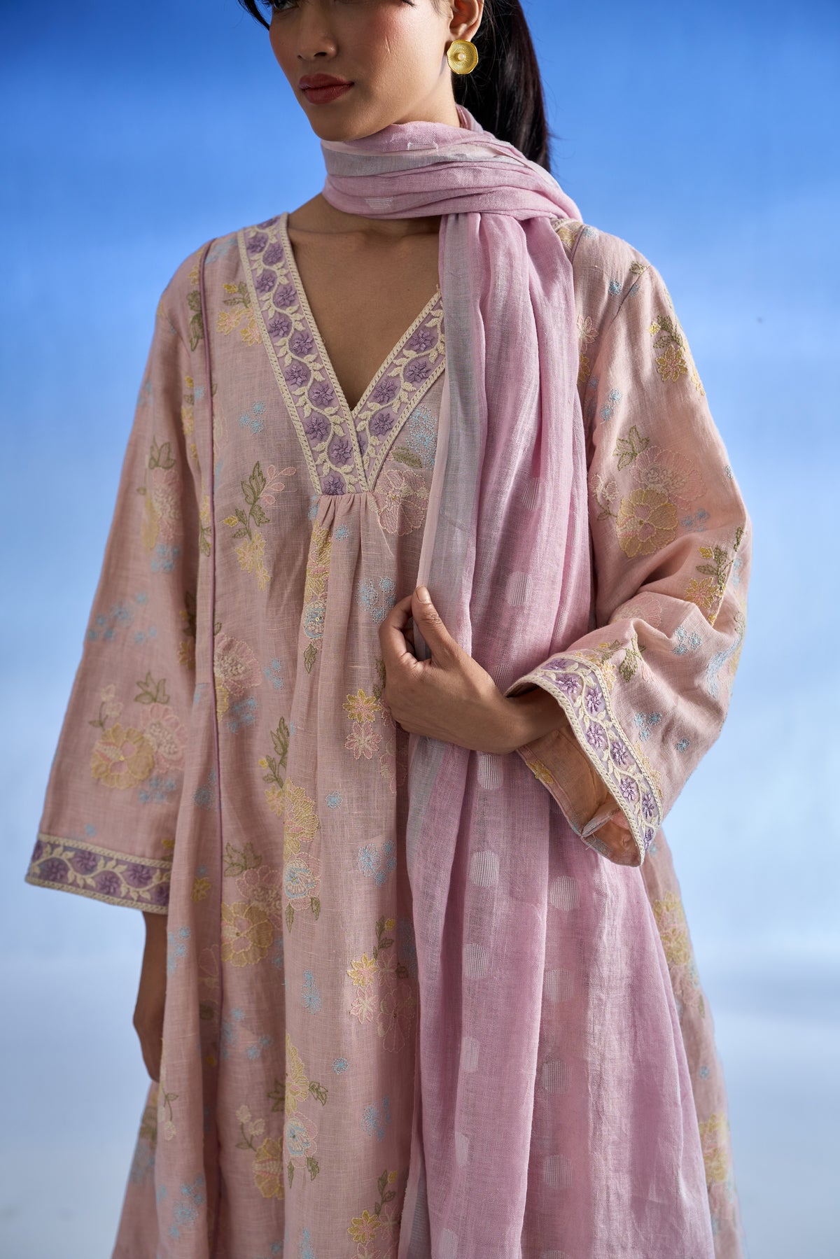 Closeup view of a woman wearing a light pink linen kurta set with floral embroidery and a matching scarf, set against a soft blue sky background.