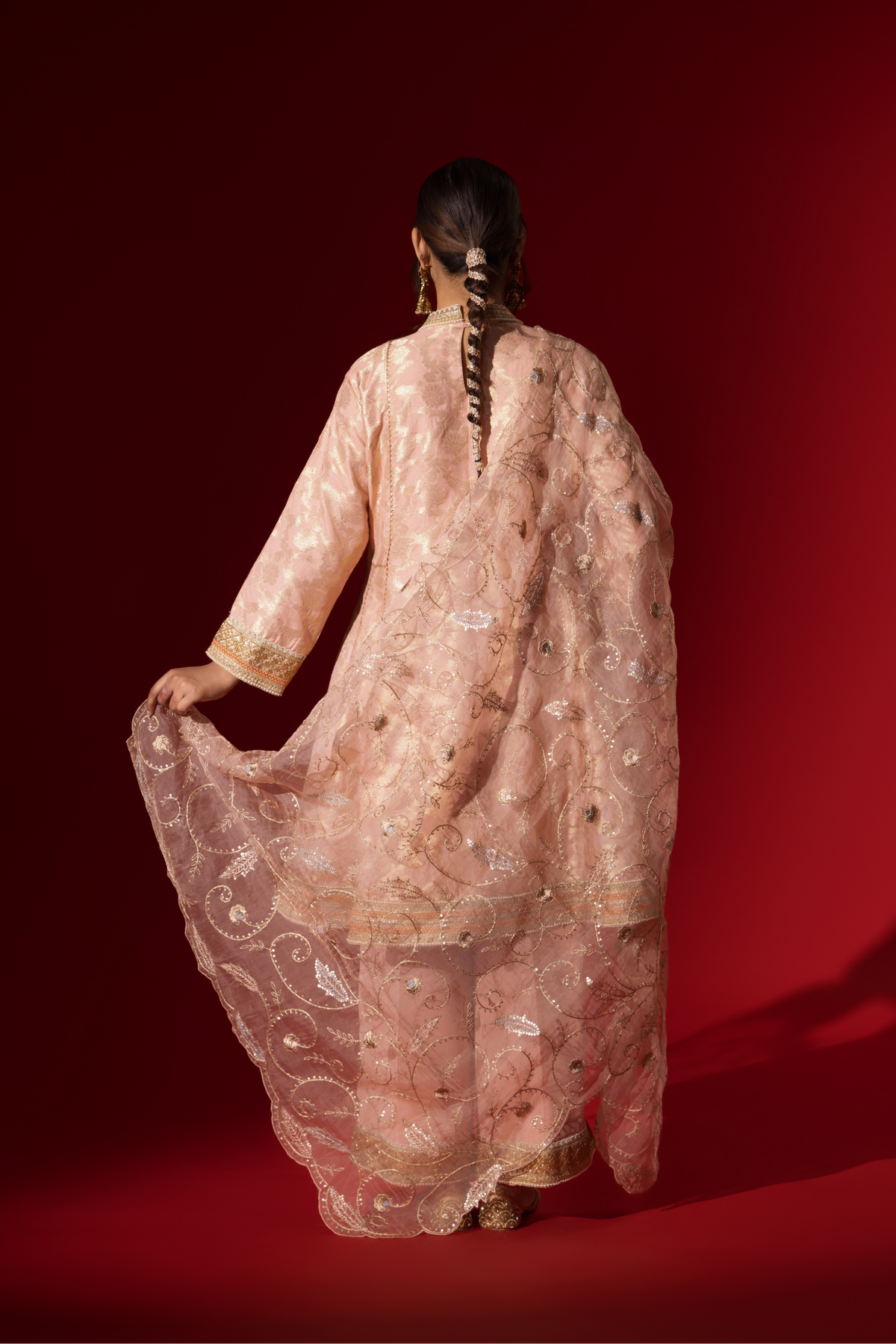 Back view of a woman wearing an elegant pink embroidered sequin brocade kurta set with gold accents and a sheer dupatta, set against a rich red background