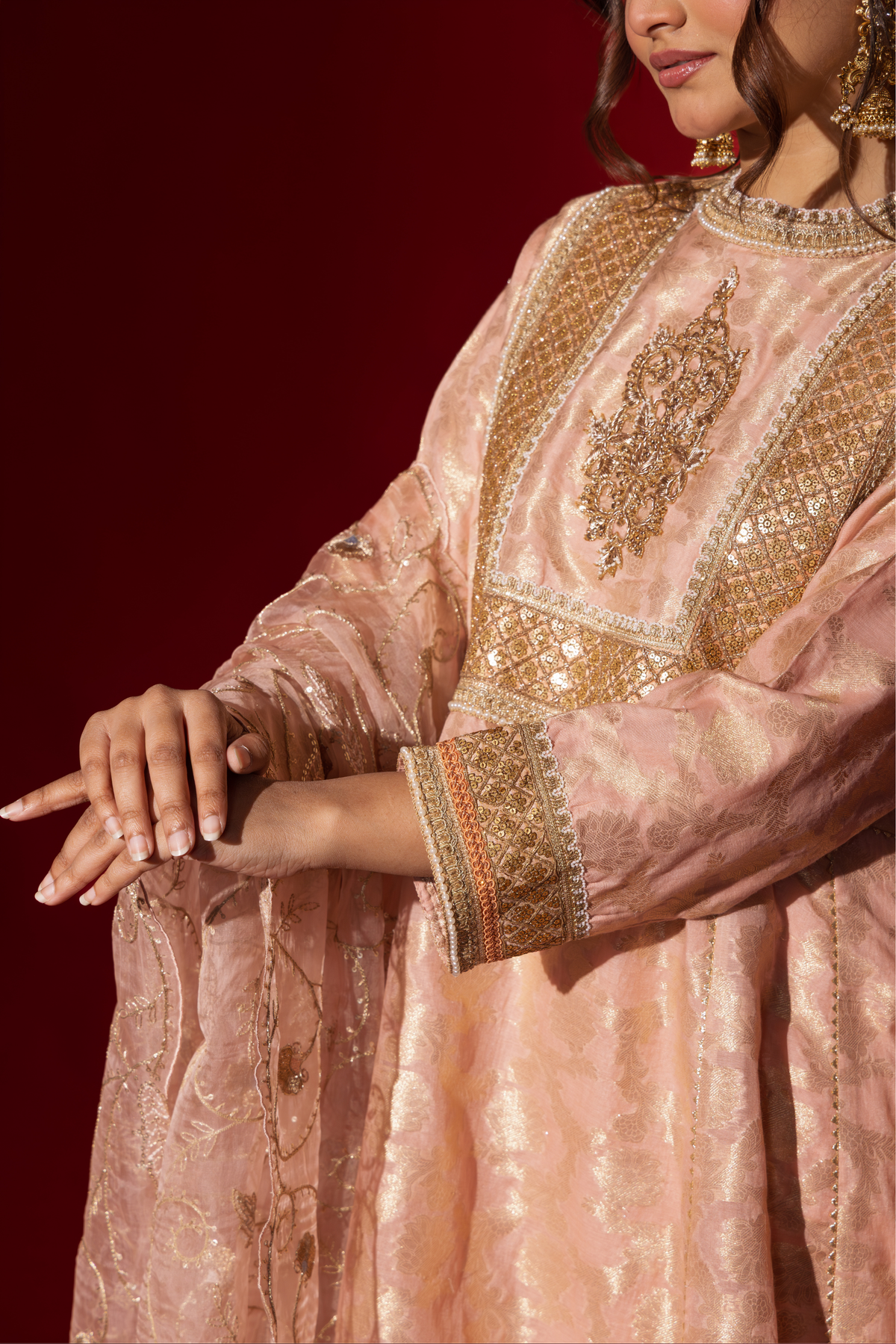 Closeup view of a woman's hand wearing an elegant pink embroidered sequin brocade kurta set with gold accents and a sheer dupatta, set against a rich red background