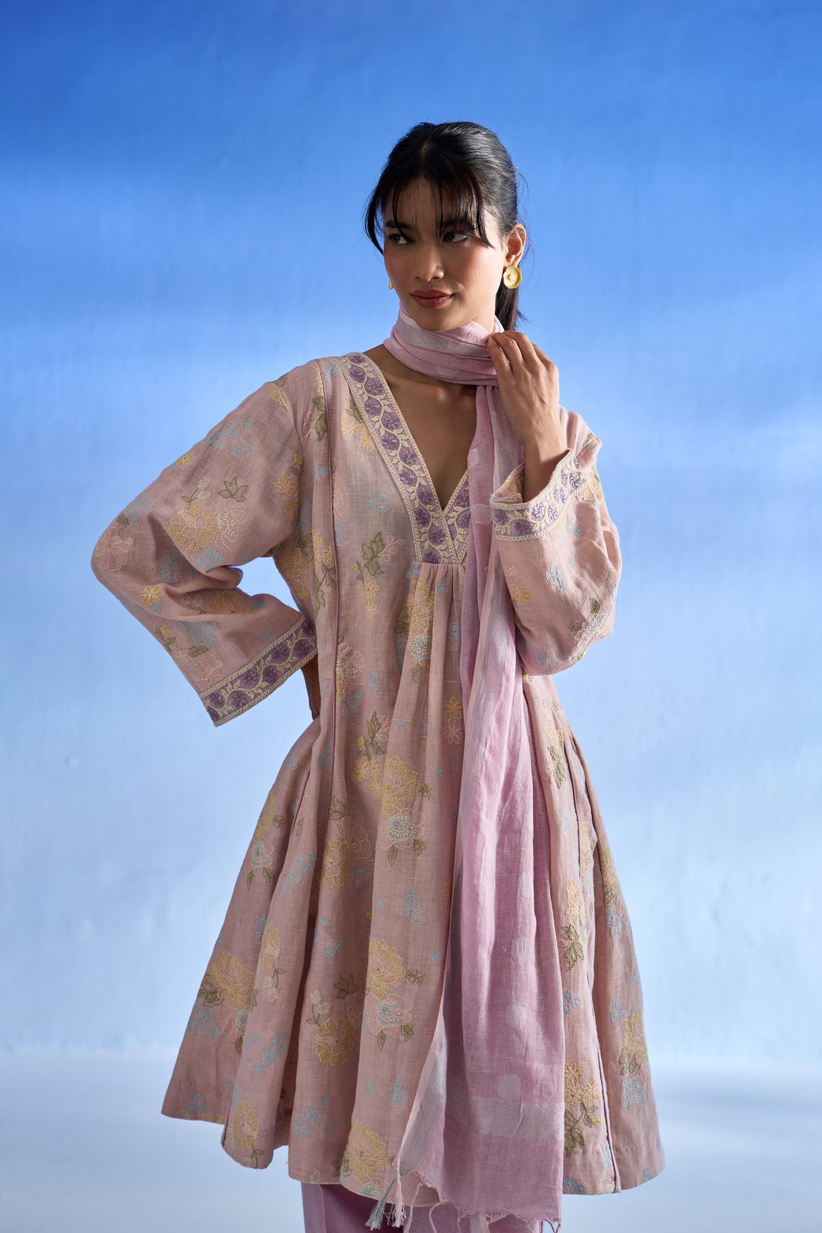 A woman modeling a rose bloom traditional outfit with a matching dupatta, featuring a floral pattern and embroidered details, set against a blue gradient background.