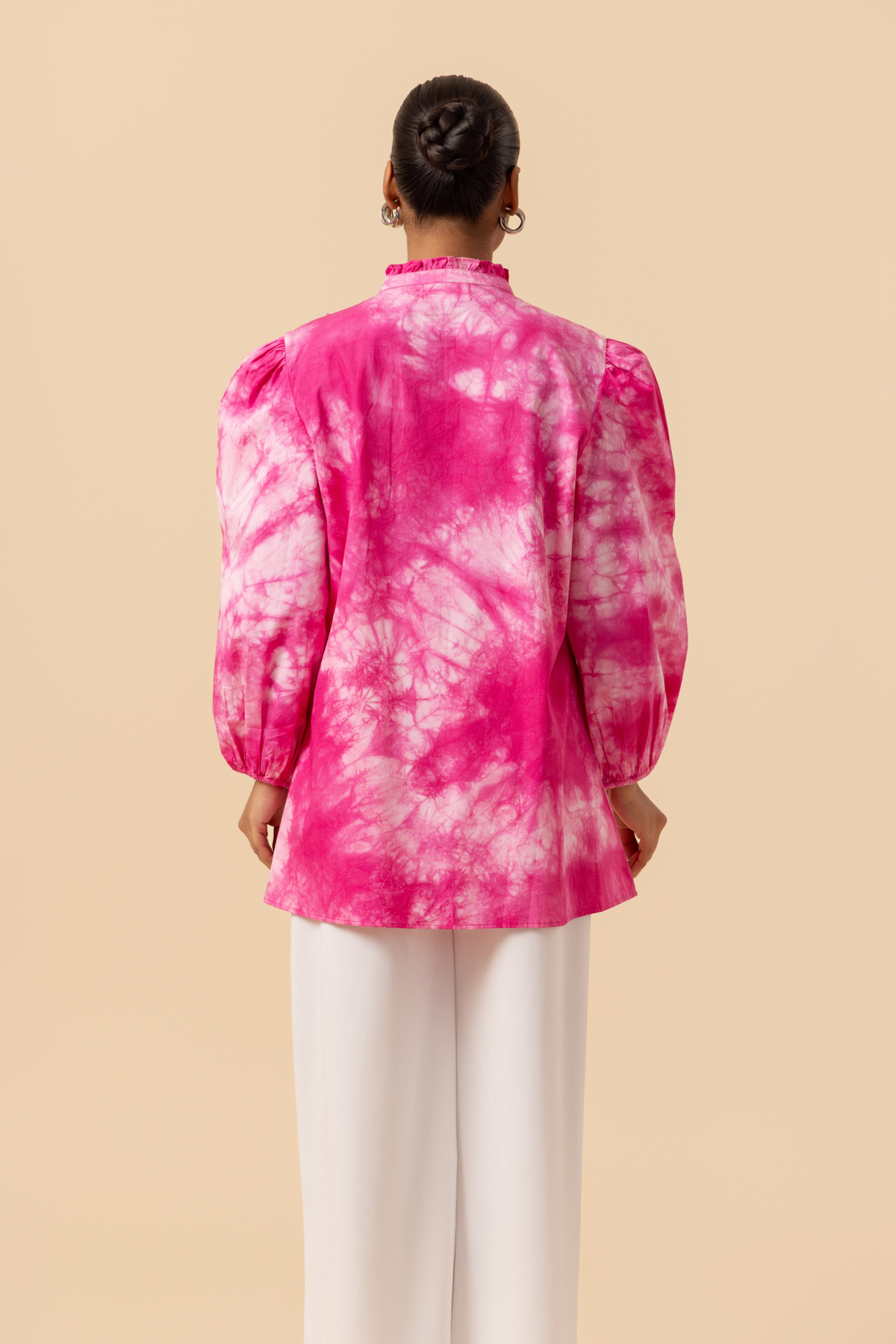 Back view of a model wears a vibrant pink tie-dye shirt dress with puffed sleeves, paired with white wide-leg trousers and stylish sandals.