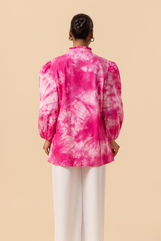 Back view of a model wears a vibrant pink tie-dye shirt dress with puffed sleeves, paired with white wide-leg trousers and stylish sandals.