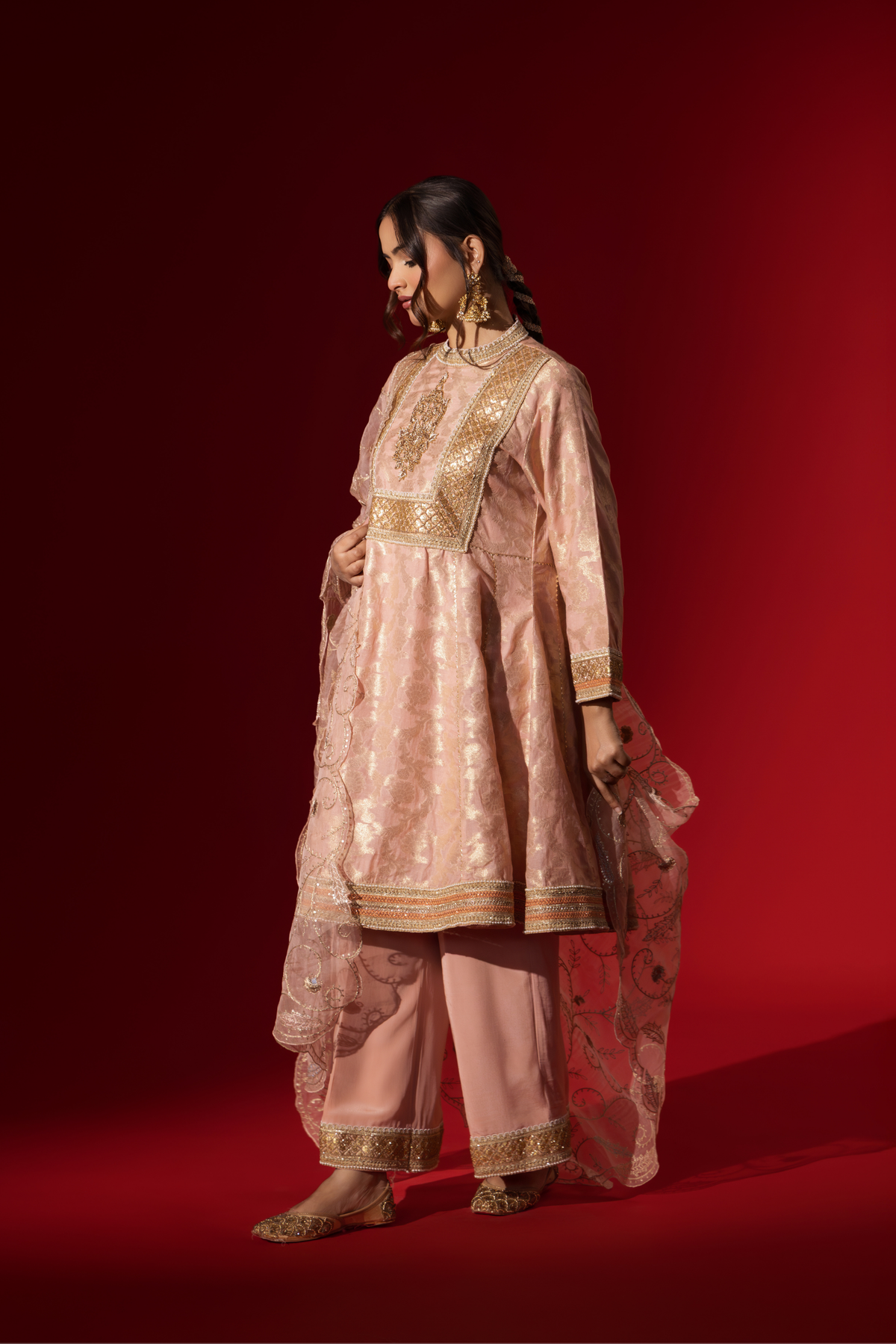 Side view of a woman wearing an elegant pink embroidered sequin brocade kurta set with gold accents and a sheer dupatta, set against a rich red background