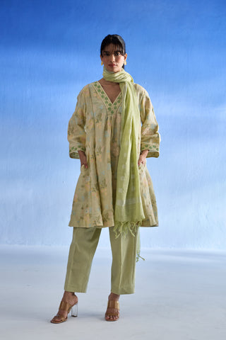 A woman models a light green and beige floral printed three-piece linen kurta set consisting of a long kurta, matching trousers, and a coordinating scarf, set against a blue gradient studio background.