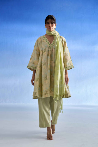 A woman modeling a pistachio green and beige floral printed kurta set with matching pants and scarf against a blue gradient studio background.