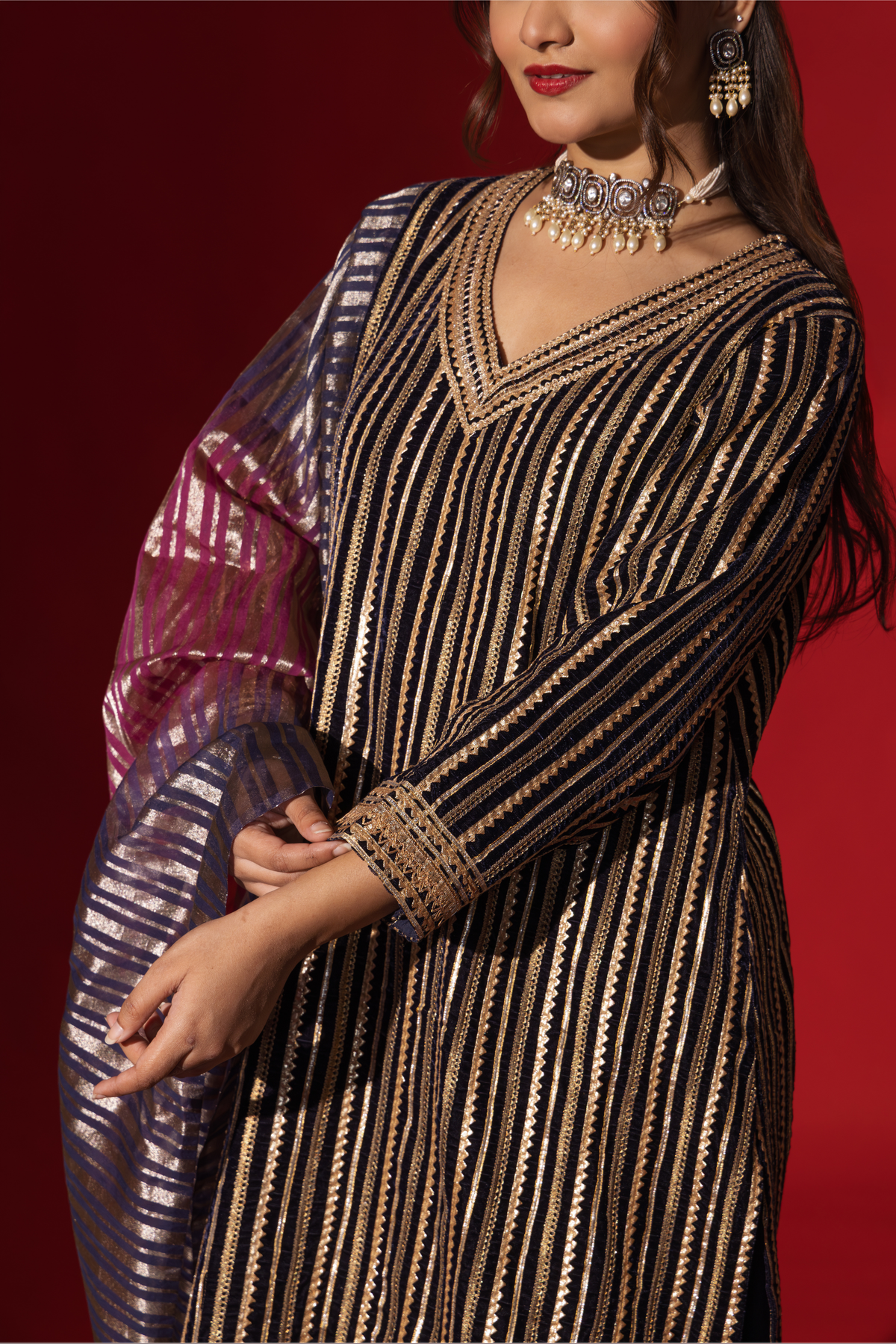 Closeup view of a woman's hand in an ethnic plum velvet gota kurta set with golden stripes, paired with a shimmering shawl, against a rich red backdrop.