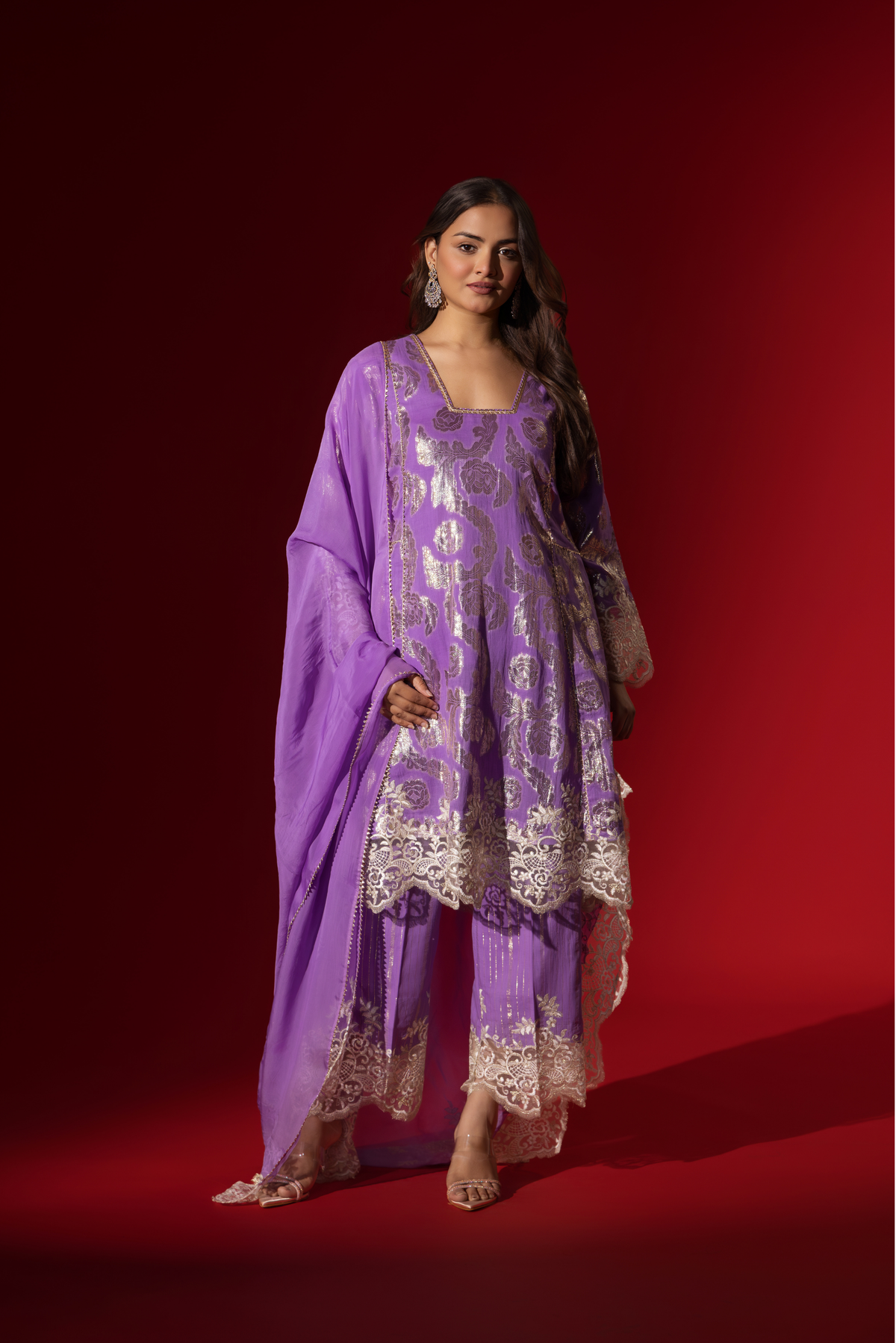 A woman poses in a purple ethnic  shimmer kurta set outfit with intricate white embroidery against a vibrant red backdrop.