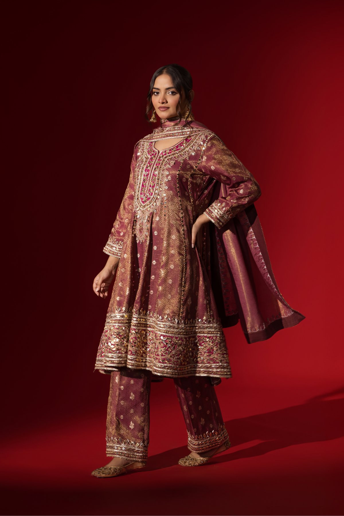 Side view of a woman stands elegantly in a rich maroon embroidered brocade kurta set adorned with intricate gold embroidery, against a deep red background.