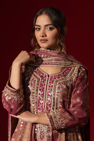 Closeup view of a woman stands elegantly in a rich maroon embroidered brocade kurta set adorned with intricate gold embroidery, against a deep red background.