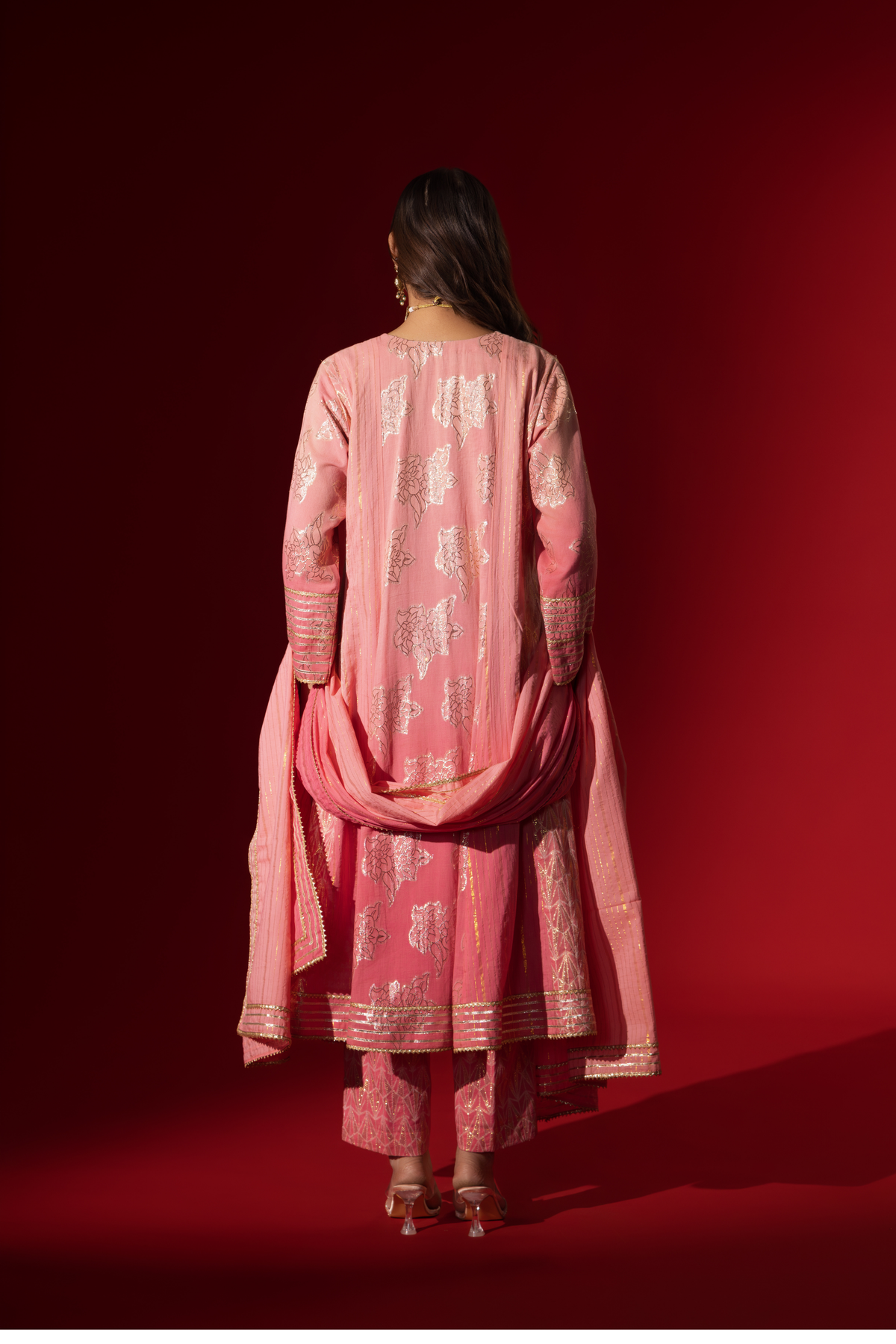 Back view of a model in a flowing pink ethnic outfit adorned with intricate golden patterns, standing against a deep red backdrop.