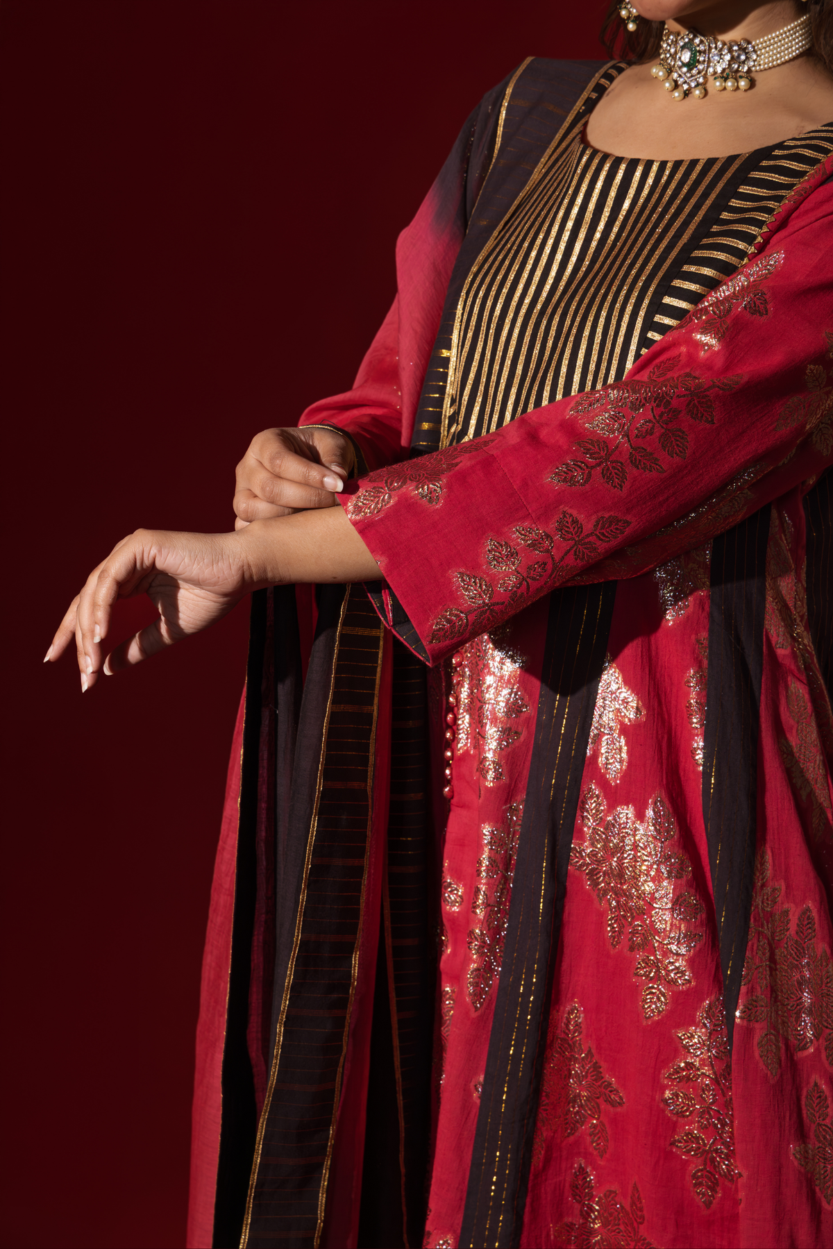 A close-up of a woman's hand gracefully posed in a vibrant red ethnic outfit adorned with gold floral designs and a black dupatta.