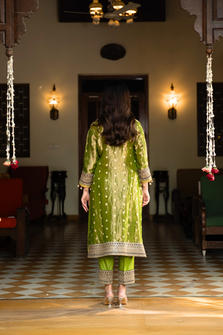 Back view of a woman in a traditional green anarkali outfit stands elegantly in a vintage-style room, surrounded by  decor and plush seating.