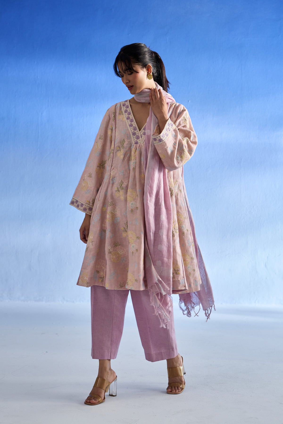 A woman modeling a light pink traditional linen kurta set with a floral print kurta, matching trousers, and a sheer dupatta against a blue gradient studio background.