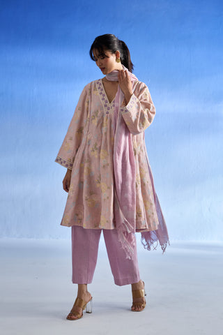 A woman modeling a light pink traditional linen kurta set with a floral print kurta, matching trousers, and a sheer dupatta against a blue gradient studio background.