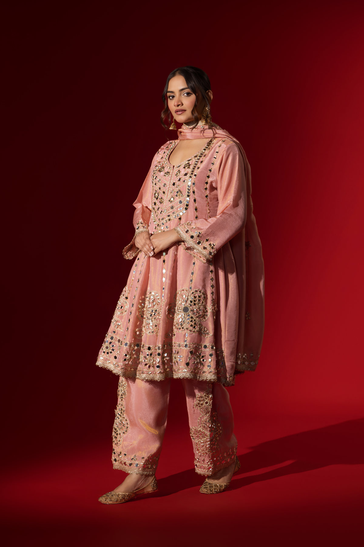 Side view of a  woman stands elegantly in a rose embroidered kurta set with intricate patterns, complemented by a matching dupatta against a bold red background.
