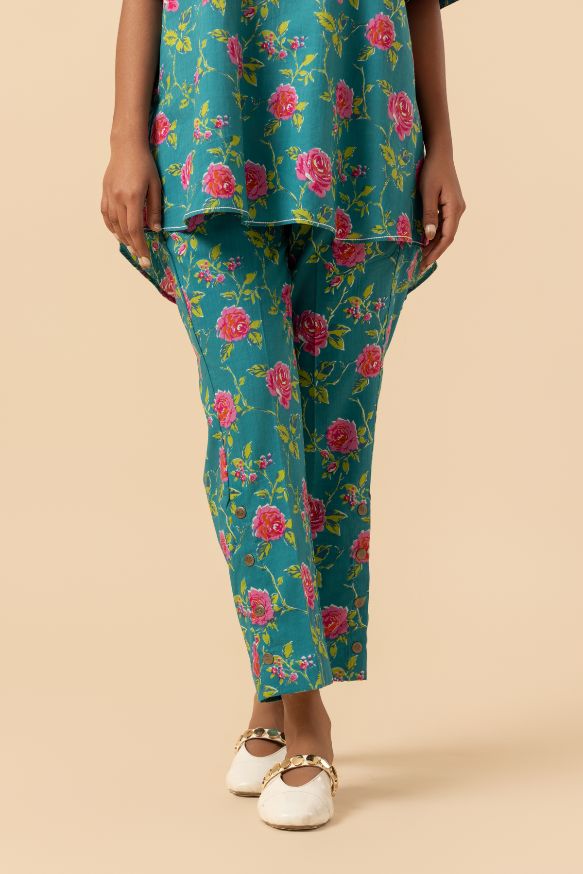 A person stands in a vibrant floral outfit, featuring a teal shirt and matching pants adorned with pink roses, set against a soft beige background against a neutral backdrop.
