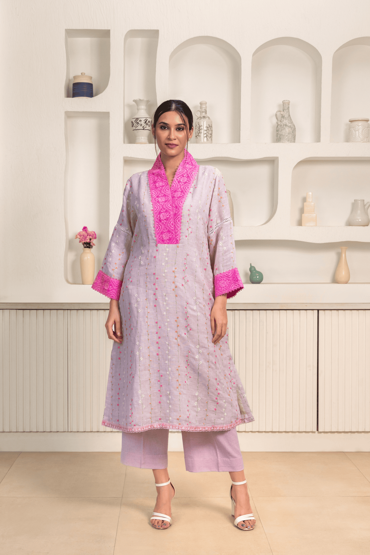 A woman is dressed in a light lavender kurta adorned with vertical embroidery patterns and bright pink lace detailing on the collar and cuffs.