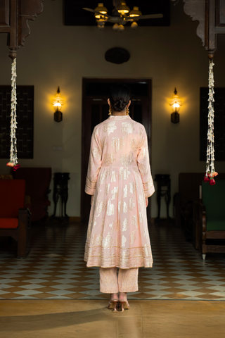 Back view of a  person stands elegantly in a pink embroidered anarkali kurta  set, surrounded by a traditional indoor setting with decorative lights and furnishings.