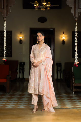 Side view of a  person stands elegantly in a pink embroidered anarkali kurta  set, surrounded by a traditional indoor setting with decorative lights and furnishings.