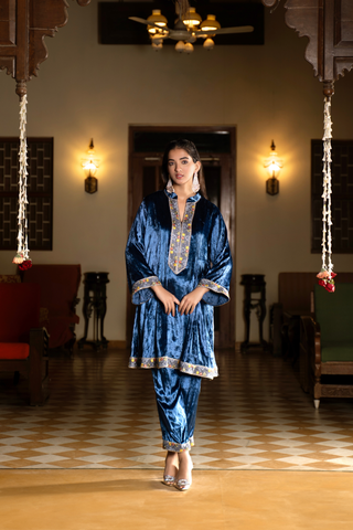 A woman stands in a traditional setting, wearing a vibrant blue velvet co-ord set outfit with decorative accents, surrounded by elegant furnishings.