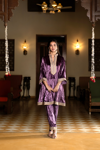 A woman stands confidently in a traditional purple velvet co- ord set outfit with intricate embroidery, set against a classic interior with warm lighting.