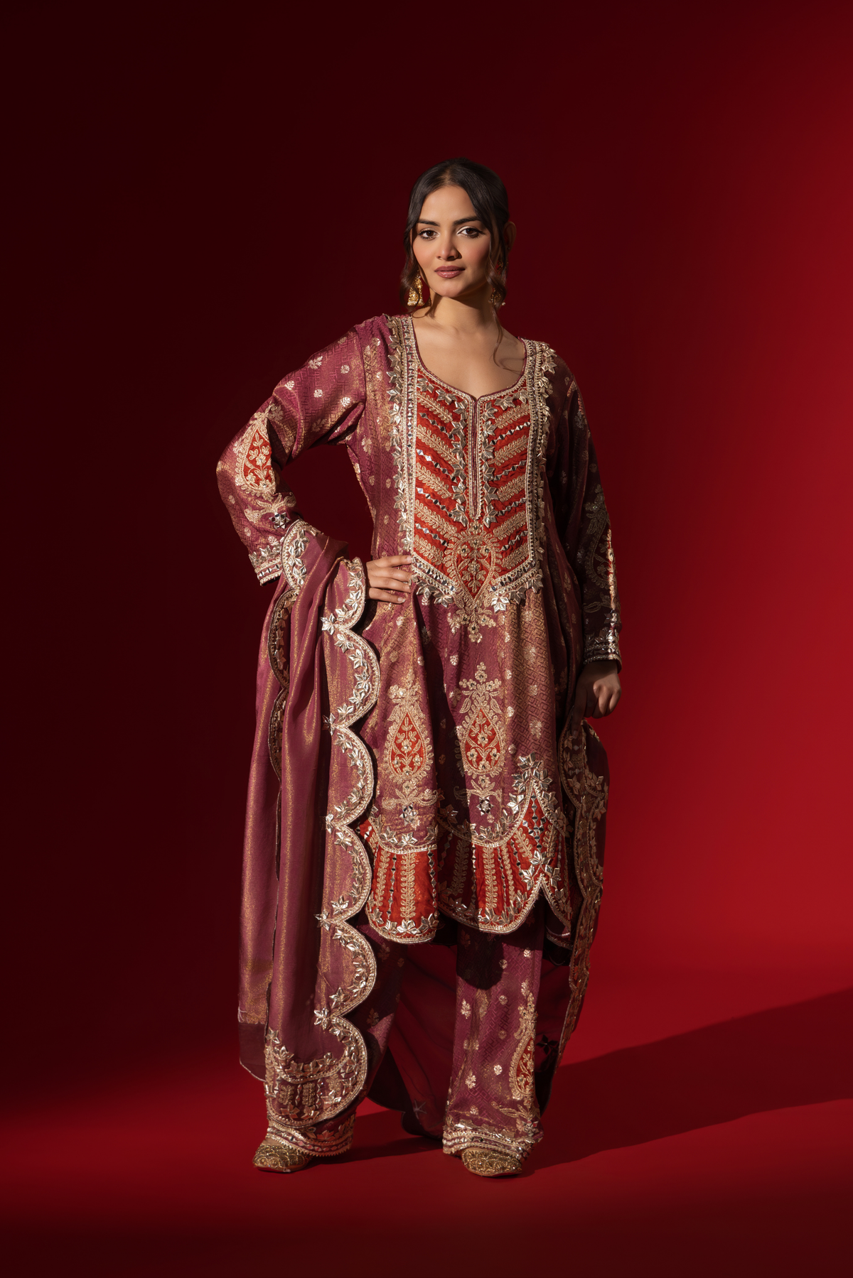 A woman wearing a richly embellished maroon embroidered brocade kurta set outfit with intricate gold detailing against a striking red background.2