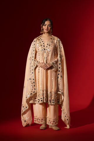 A woman stands elegantly in a beige embroidered kurta set with intricate patterns, complemented by a matching dupatta against a bold red background.