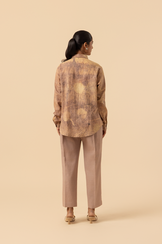Back view of a model stands against a light beige background, wearing a patterned co-ord set which has button-up shirt and wide-legged beige pants, accessorized with heels.
